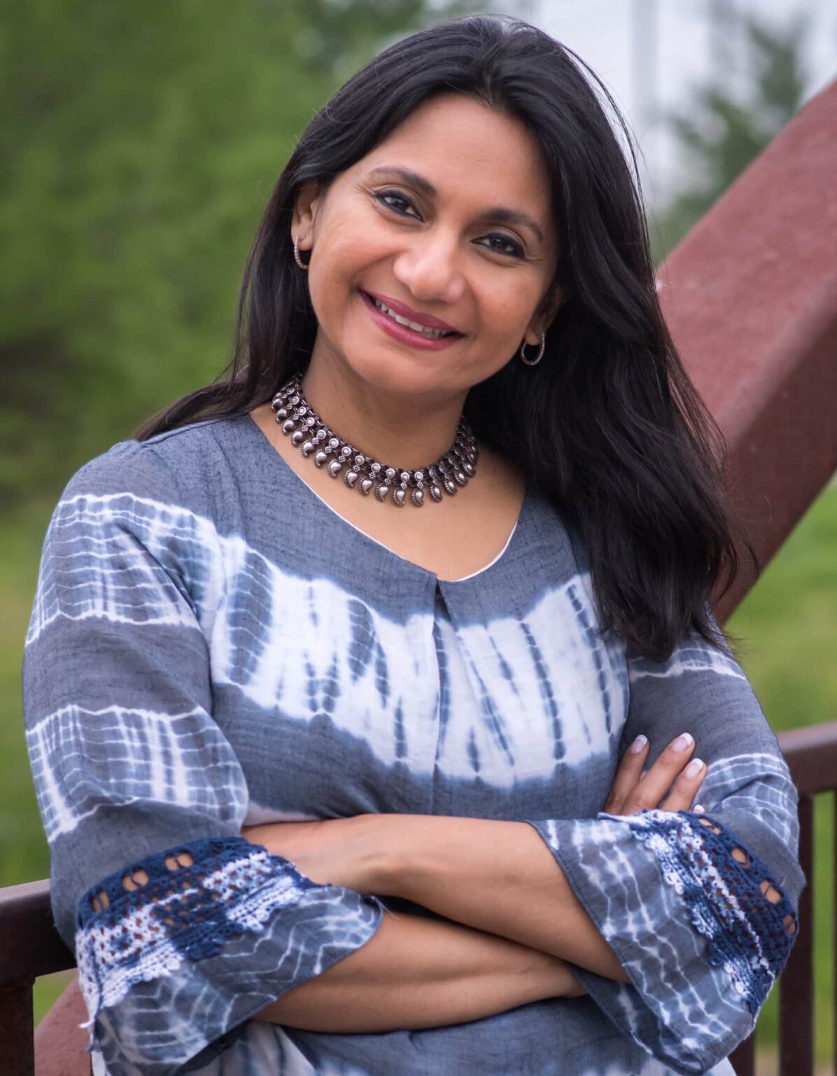 woman in dark hair, necklace, and blue-gray and white shirt smiling with arms crossed
