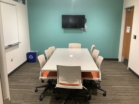 Photo of the Conference Room, with a table, eight chairs, a monitor on the wall at one end of the table, and white board on the left wall.