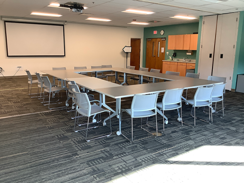 Photo of the full Meeting Room, with tables, chairs, a projector screen, and small counter and sink.