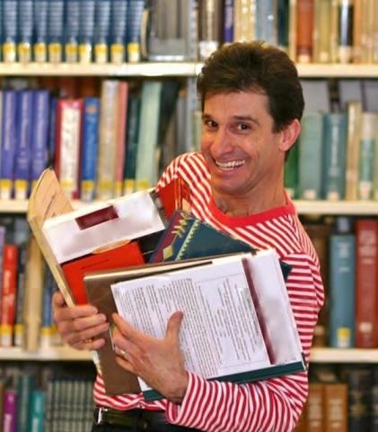 Chris Fascione wearing a red and white striped shirt, holding books and standing in front of shelves of books