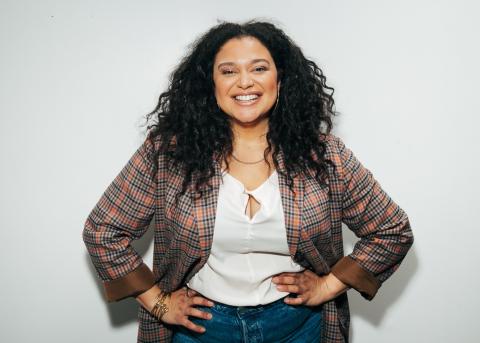 Michelle Buteau smiles into the camera while set against a white back drop.  She is wearing jeans, a white blouse buttoned at the bodice and a plaid blazer with the sleeves folded to the third quarter.  Her hair is loose and hangs in curls about her face. 