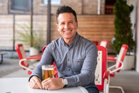 Steve Dolinsky sits at a patio table, holding a tall glass of beer that is amber in color with a foamy top.  He is wearing a gray long sleeve shirt, buttoned to the neck and smiles at the camera. 