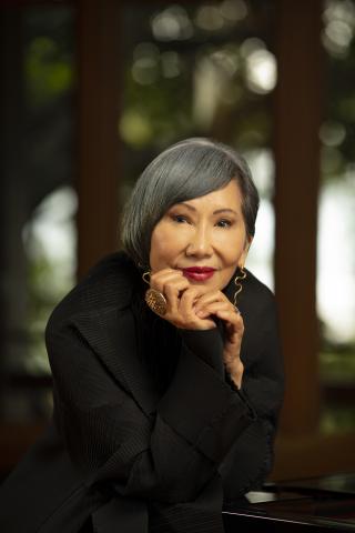 Amy Tan stares into the camera with her chin in her right hand. Her salt and pepper hair is parted to the side and falls over her right shoulder.  She is wearing all black and the background is blurred in shades of black and grays