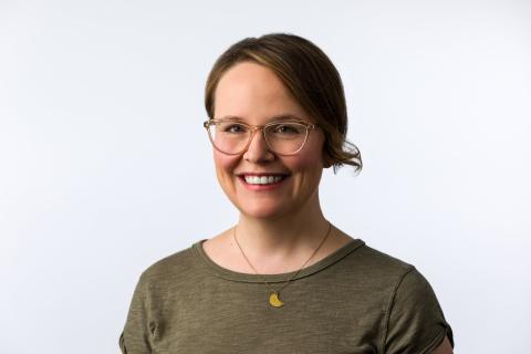 Raina Telgmeier against a white back ground.  She has on an olive green top with a gold half moon necklace.  She is wearing her hair in a low chignon and has on clear acrylic glasses.