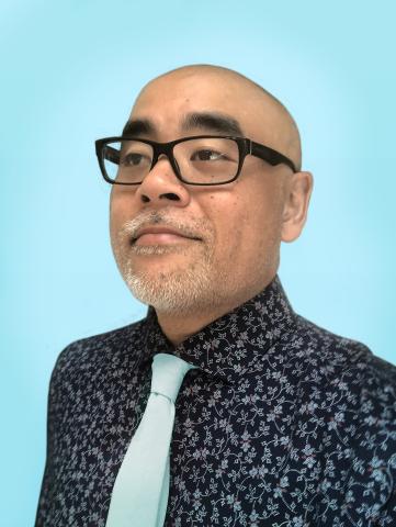 A man of color stands looks off camera, set against a sky blue background.  He is wearing a black shirt with a bright pattern and a light blue tie, and a pair of plastic black glasses.  