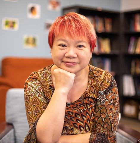 Asian American professor Dr. Ada Cheng sits with her hand placed on their right fist.  Their hair is strawberry and they are wearing a patterns shirt. 