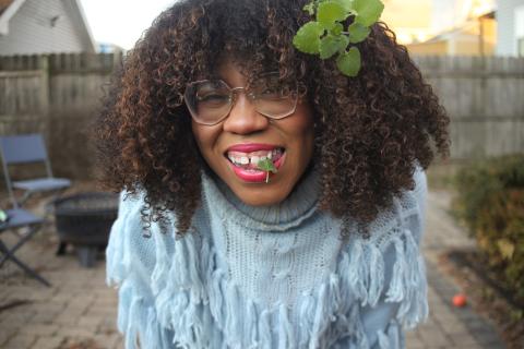 A smiling African American woman is front and center, dressed in a baby blue sweater, a bright red lipstick and a smile, as she is standing in a back yard with a wooden fence surrounding