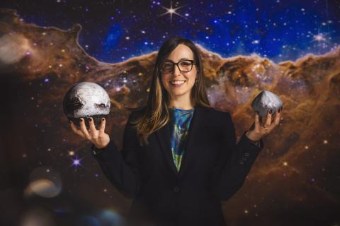 Standing against a backdrop of space, a white woman with glasses and long hair, parted and laying on her right shoulder, holds her hands up.  In one hand she holds a model of Earth and in the other, a model of a dense gray asteroid. 