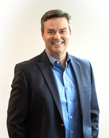 Author Gary Freund stands against a cream background in a blue shirt and jacket, smiling confidently at the camera