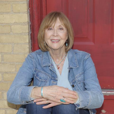 A middle aged white woman sits on the stoop of a building, with a red door and brick behind her.  She is wearing a light colored denim jacket, casual light blue shirt, and a few pieces of silver jewelry.  Her hair is a light red-blonde and is cut to her shoulders. 