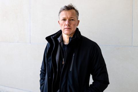 A middle aged white man stands against a grey-white background.  He is wearing a black shirt with a black leather jacket over it. His color is popped around his neck.  He has short salt and pepper colored hair 