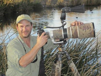 Set against the back drop of a marshy lake, a man stands facing the camera, with his hands on his own beige camera, mounted to a beige tripod.  The man is wearing a tan colored hat, a short sleeved neutral shirt. 