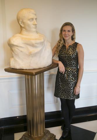 Mallory Mortillaro stands next to the Rodin Bust she re-discovered.  She is wearing a black and gold dress with a black headband in her long blonde hair.  The bust is large, and a creamy ivory color,  It is a of a regal looking man.