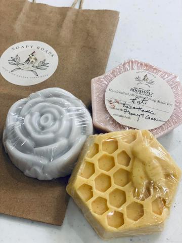 an image of three handmade soap bars on a table with a brown paper bag,  one looks like a honeycomb, one looks like a gray rose, and the other is a pink octagon shape