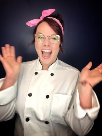 Chef Lauren Ocello exuberantly looks at the camera. She is wearing a white chef coat, her hair is back in a pink kerchief, tied into a bow.  Her hands are up in mid-clap, 