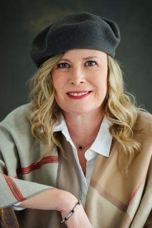 Headshot of Donna Kathryn Kelly wearing a black beret over her long blond hair.  She is wearing a beige, taupe and red sweater over a crisp white shirt. 