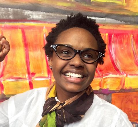 A young African American woman stares at the sky behind a orange and yellow backdrop.  She is dressed in vintage clothing, with her hair in short curls, and black acrylic glasses