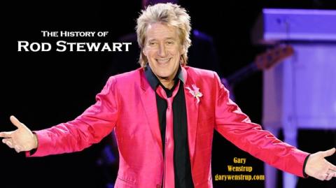 A spunky older man dressed in a hot pink blazer, black shirt, and a hot pink tie stands open armed on a stage