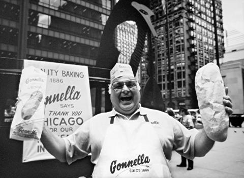 Black and White photo of a Baker from Gonnella's bakery.  He is wearing a bakery hat, apron and short sleeved shirt.  He is holding a loaf of bread in his left hand and a Gonnella bakery bag in his right hand