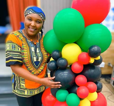 Educator Elizabeth Kahura stands holding a colorful bouquet of balloons in green, yellow, black, and red.  She is wearing a traditional African style top and cap.  