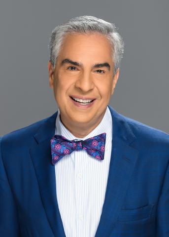 Paul Lisnek is featured on a gray background.  He has white pepper hair cut short above his ears and neatly combed to the right.  He is wearing a navy suit, with a white button up shirt, with a with a blue and fuschia bow tie. 