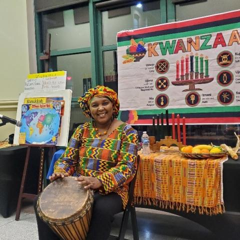 A cheerful African woman stares at the camera dressed in traditional regalia.  The colors are a deep yellow and green.  She is holding a traditional hand drum.  