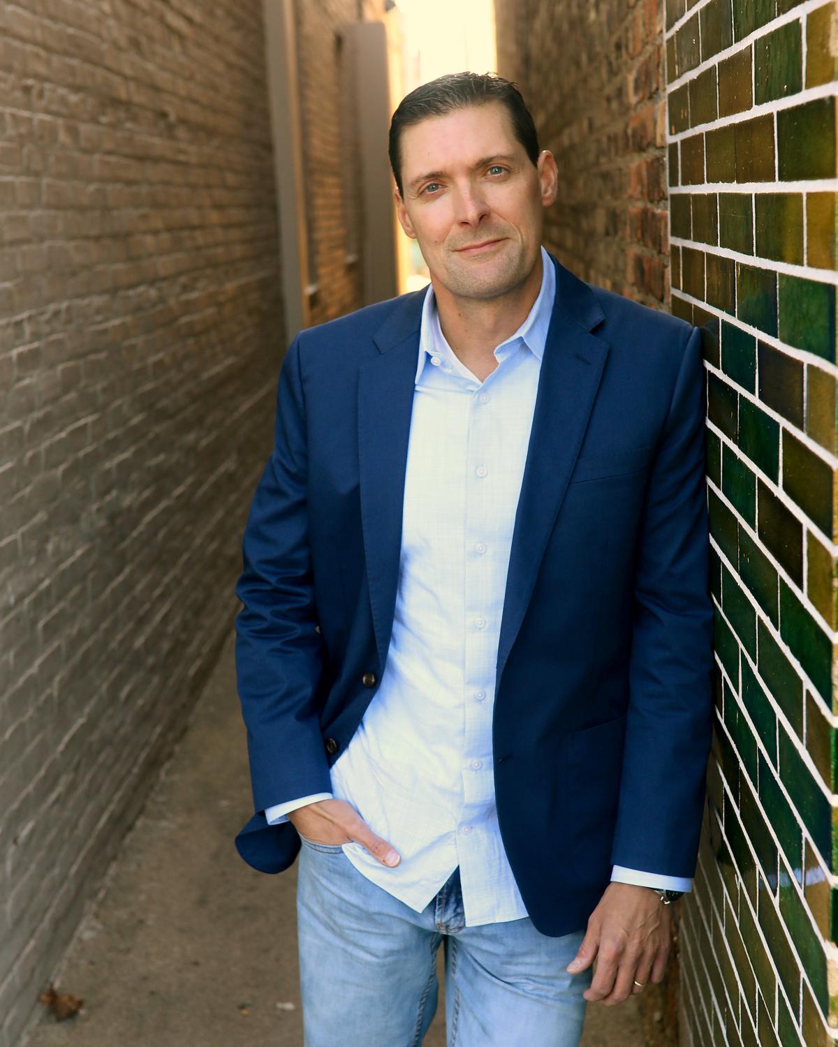 dark-haired man standing in well-lit alley wearing button-down shirt, sportscoat, and jeans