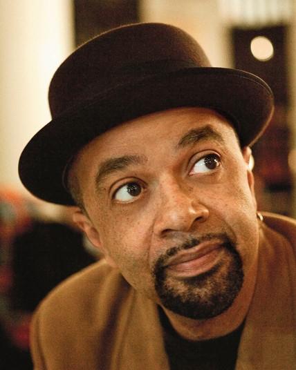 James McBride is shown against a blurred sepia background.  He's wearing a darn bowler cap, a brown overcoat with a black crew neck shirt beneath it.  He eyes peer up and into the right corner of still and his goatee is neat and clean around his pursed lips. 